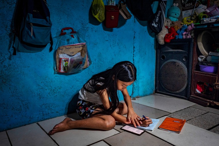 A 12 year old girl sitting on a tiled floored in a home doing school work, using her mobile phone and writting in a notebook.