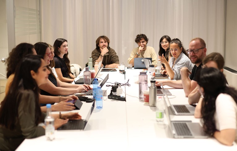 A large group of people sit around a long rectangular white table with laptop computers and bottles of water as Manuel Spitschan holds a lab meeting.