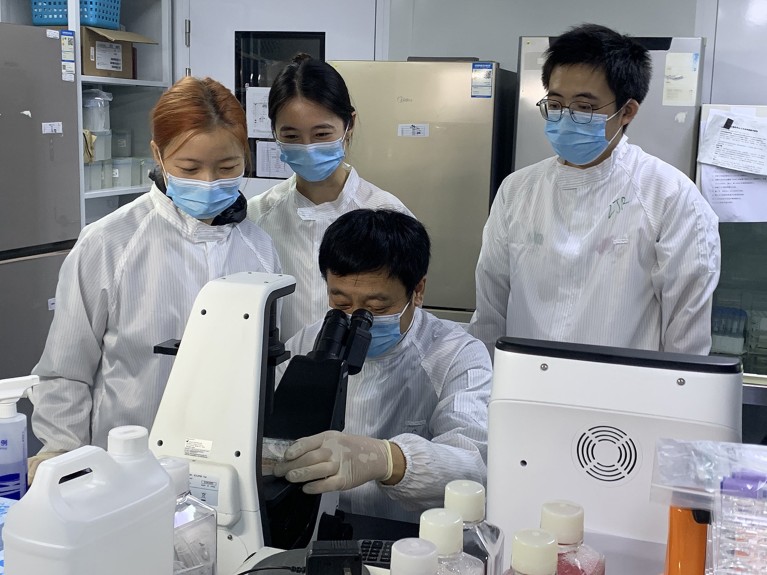 Hongkui Deng operates a microscope in a laboratory, while three researchers wearing face masks and cleanroom lab coats gather around him.