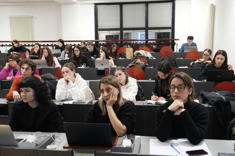 Students sitting in a tiered auditorium work on laptops and take notes.