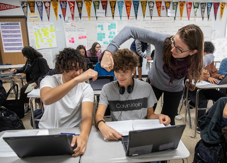 Seatec high-school student with a laptop receives congratulatory fist bump from teacher. Classroom walls display university pennants, posters and notes.
