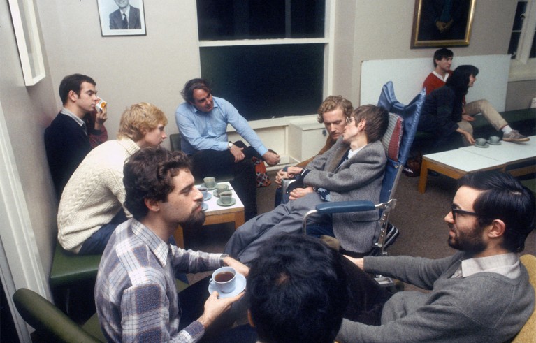 Stephen Hawking talks to graduate students seated around a small table with teacups