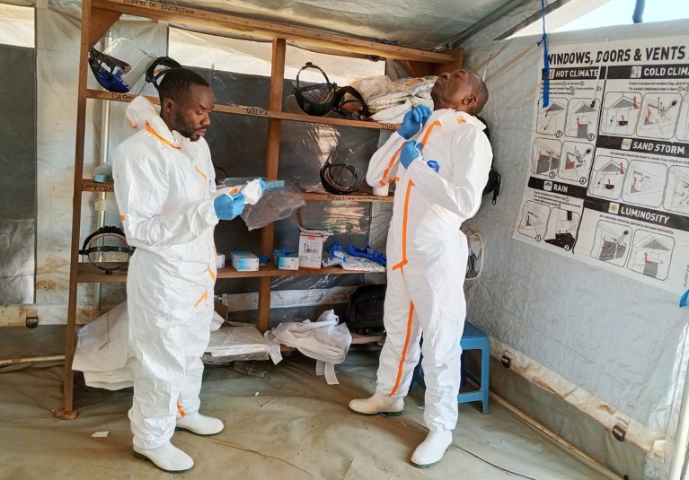 Richard Kojan zips up his white personal protective equipment suit as he stands with his colleague Patient Kipala Wenga, who is about to secure his own face mask.