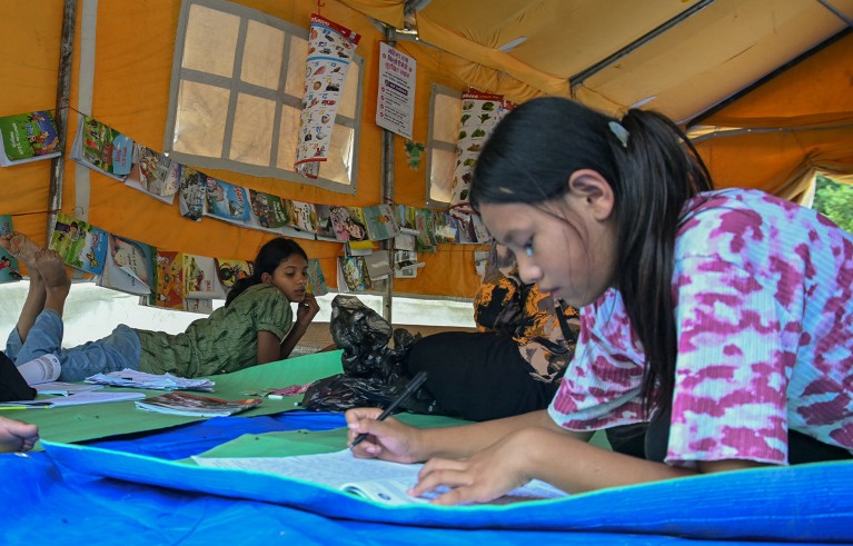 Children studying while lying on floor mats in a tent, amid colourful educational displays and posters.