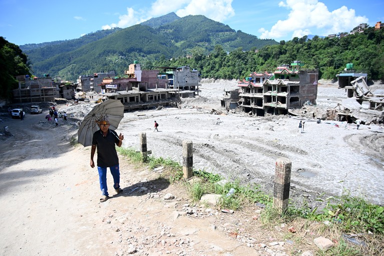 A man carrying a sun umbrella walks past muddy houses damaged by flash floods in Nepal, with green mountains rising in the background.