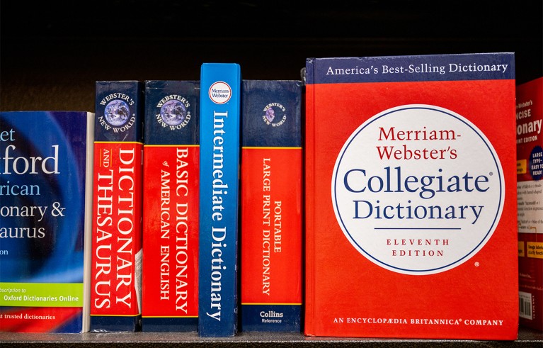 Upright dictionary books on a shelf, representing English-language reference works for definitions and usage. Visible titles include Oxford, Collins and Merriam-Webster dictionaries, with red, blue and navy covers, including Merriam-Webster’s Collegiate Dictionary.