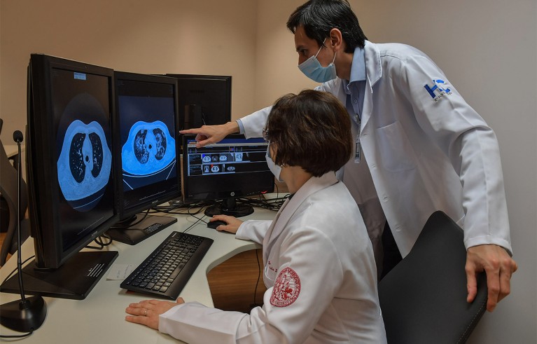 Two masked radiology doctors with white coats review lung CT scans on computer monitors.