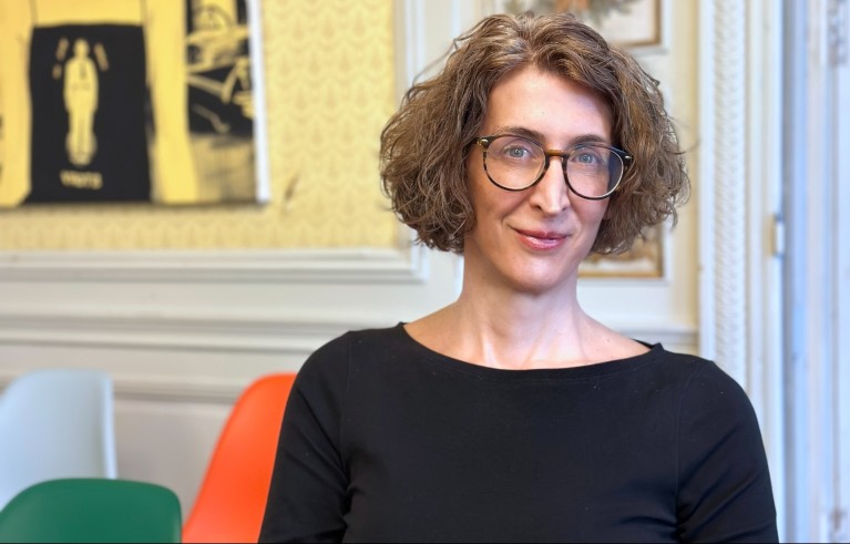 A portrait of Mirta Galesic against a background containing framed artwork, patterned wallpaper, white panelling and colourful chairs.