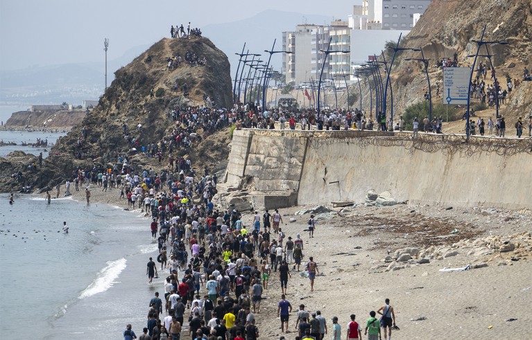 Migrants crowding a beach and coastal border area at Ceuta, Spain. Headline reports more than 48,300 migrants returned to Morocco, with fences, rocky cliffs, dense crowds.
