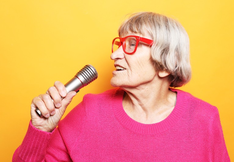 Older adult speaking into a handheld microphone with a bright background.