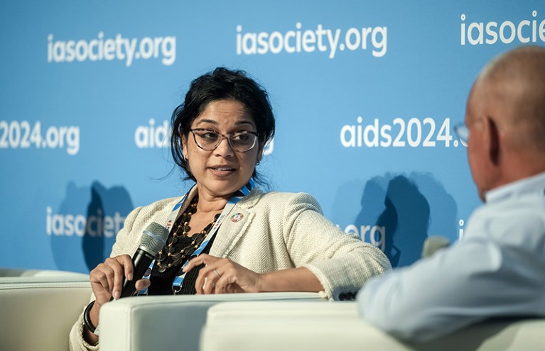 أنجيلي أشريكار يتحدث في إحدى جلسات مؤتمر aids.2024.org في ميونيخ، ألمانيا. تحمل ميكروفونًا وهي تجيب على سؤال. اللافتة الموجودة خلفها تقول iasociety.org وids2024.org.