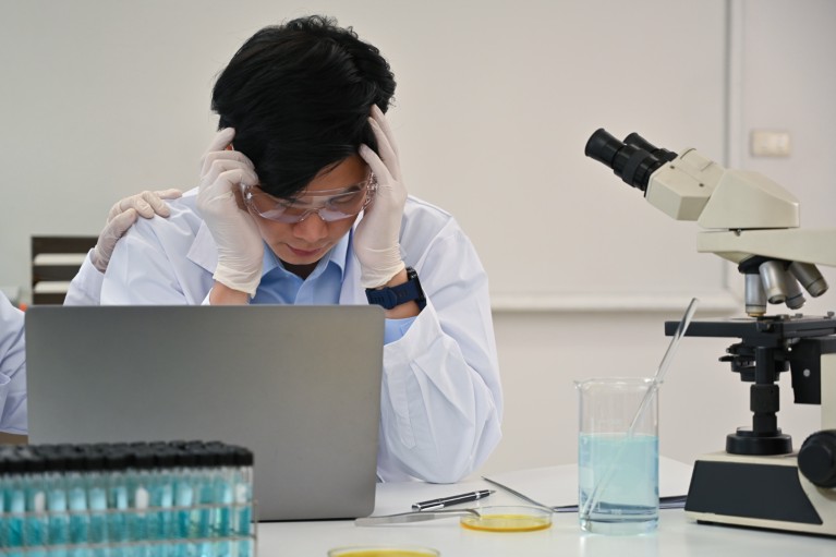 A laboratory worker in protective gear and a lab coat sits at a laptop beside a microscope and test tubes with his head in his hands