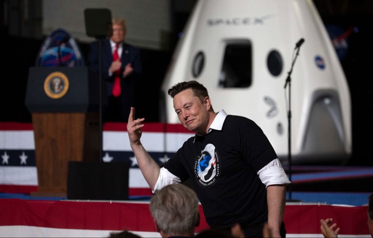 إيلون ماسك يتحدث في إحدى فعاليات SpaceX، وخلفه كبسولة مركبة فضائية ودونالد ترامب على منصة تحمل الختم الرئاسي.