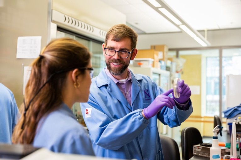 Michael Jewett smiling and gesturing at a Petri dish in a laboratory while talking to a student.