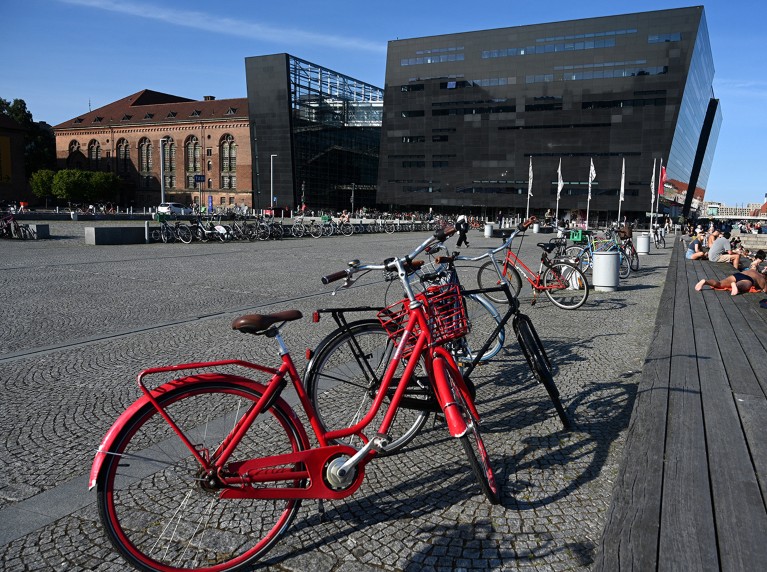 An old-fashioned red bicycle is seen in the foreground of a riverfront promenade in Copenhagen, with the National Library of Denmark in the background.
