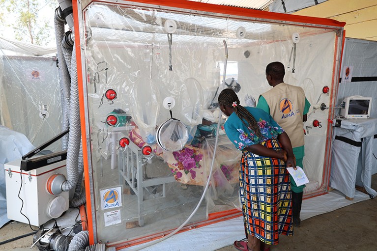 A patient is tended to by two nurses as they lie on a bed in a CUBE. A CUBE is a square medical quarantine structure with transparent plastic walls that feature integrated glove sleeves, allowing doctors and nurses to provide care, monitor vital signs and talk to people diagnosed with Ebola from outside it's walls.