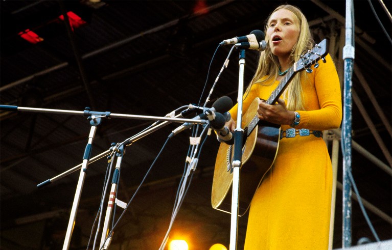 Joni Mitchell, a blonde female musician singing into a microphone while playing acoustic guitar during a live outdoor performance. She wears a bright yellow dress, with multiple microphone stands, stage lighting and metal rigging surrounding her.