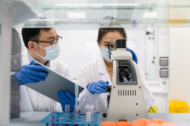 Two researchers work in a lab at a microscope using a digital tablet.