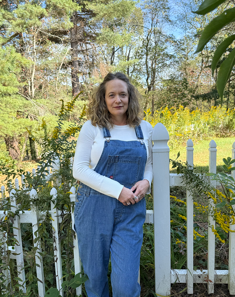 Rebecca Lindsay leans on a white picket fence in a lush green garden.