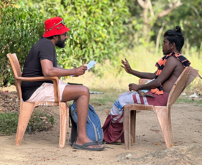 Eranga Galappaththi sits on a chair outside, talking to a village leader, also sitting in a chair..