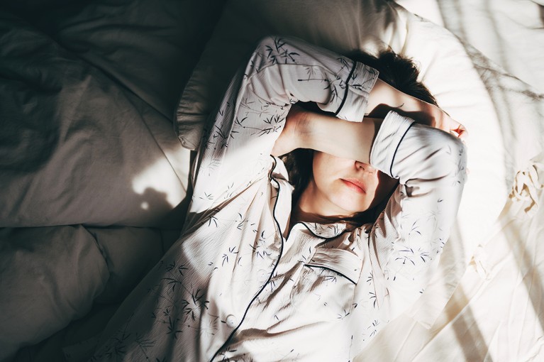Overhead view of a young woman laying in a bed covering her eyes with her arms, as the sunlight floods into the room.