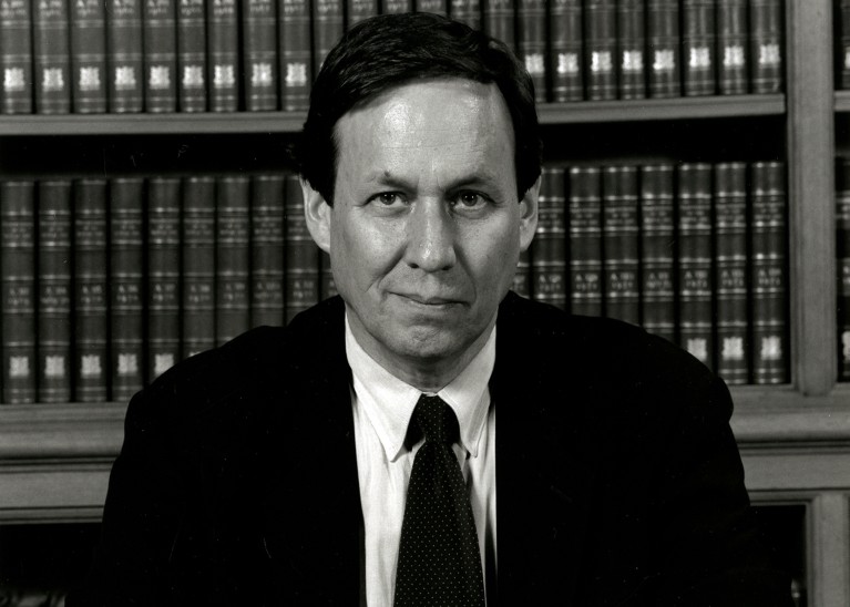 Portrait of Robert Williamson looking into the camera while sitting at a table in front of a bookshelf.