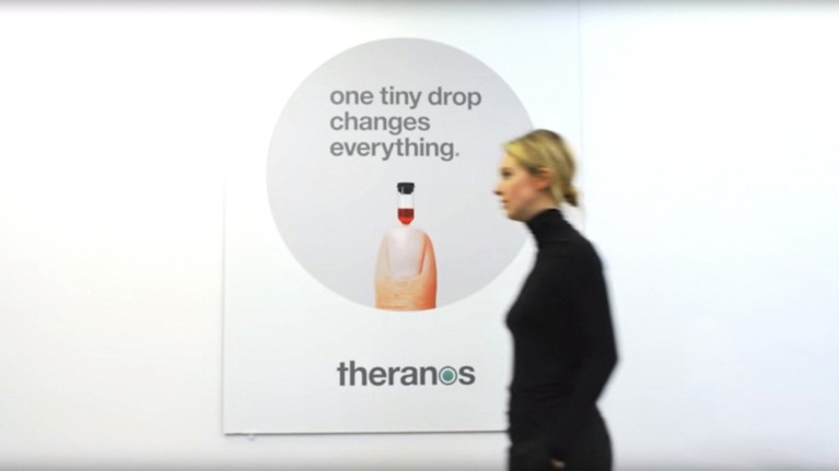 Movie still with Elizabeth Holmes walking in front of a poster promoting Theranos, from “The Inventor: Out for Blood in Silicon Valley”, 2019.