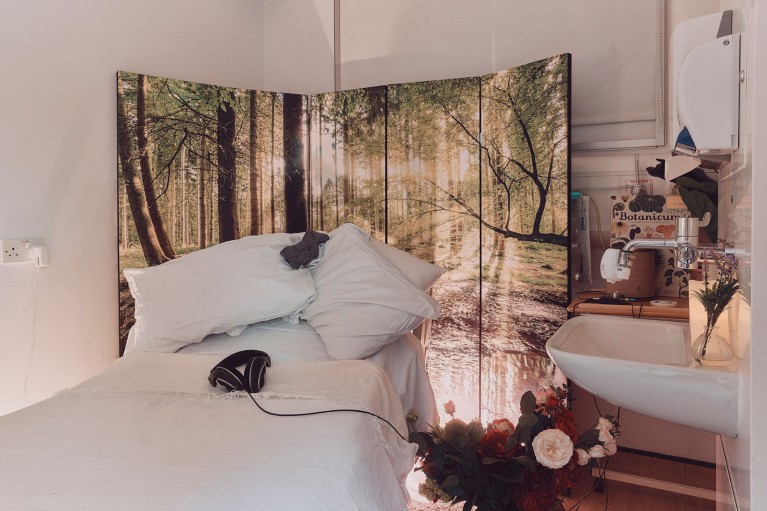 A room with a white bed and headphones placed in front of a folding screen with a sunlit forest scene printed on it, next to a sink with a vase of flowers.