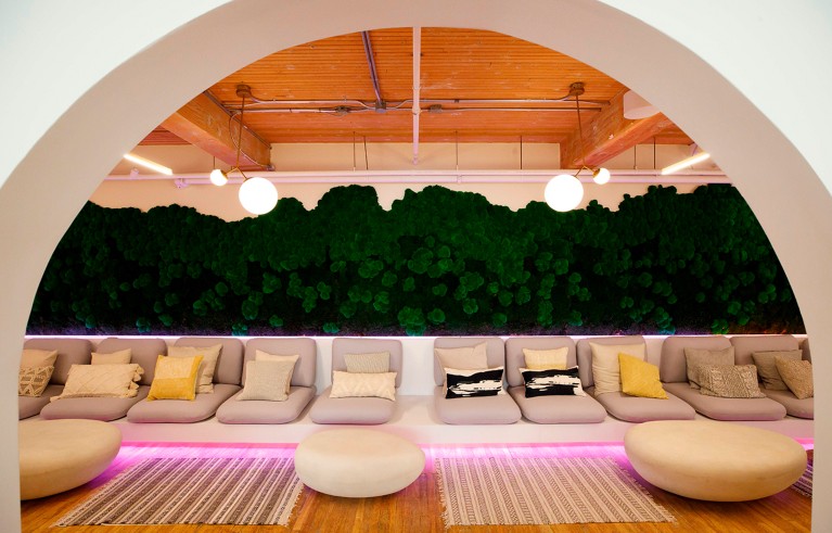 A warmly lit room with beige seating and patterned rugs in front of a green plant wall.