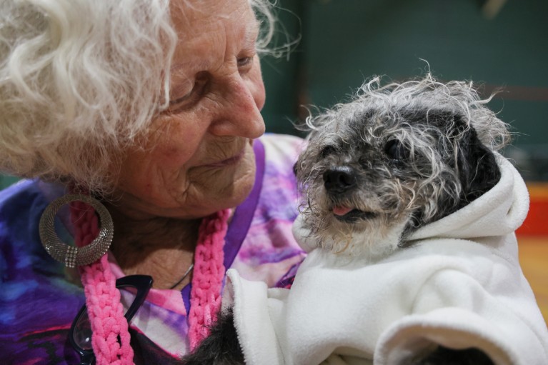 Close-up of a woman with white curly hair holding a small black-and-grey dog wearing a white hooded jumper