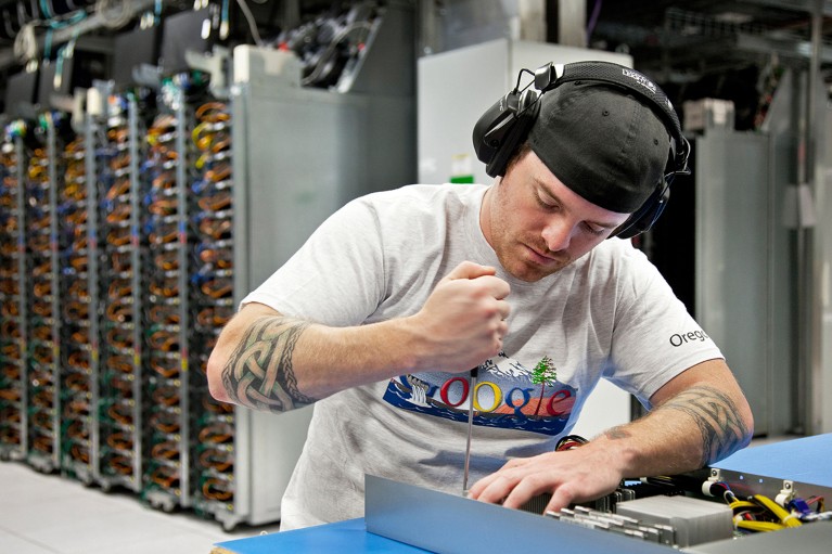 A man wearing a hat, headphones, and a google t-shirt leans over a piece of work on a bench, using a screwdriver.