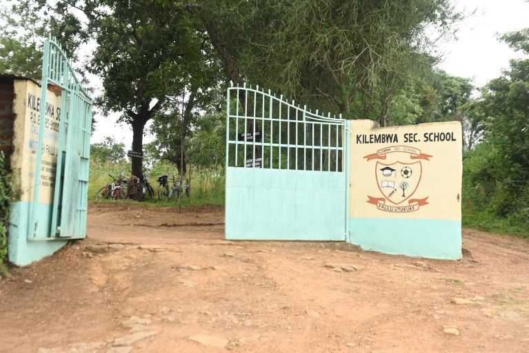 A dusty dirt track leads through gates, displaying the coat of arms for the Kilembwa secondary school