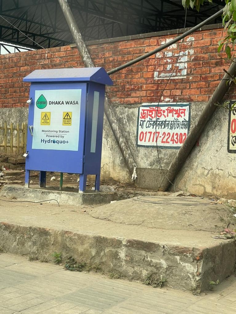 A roadside cupboard is labelled as a monitoring station for Dhaka WASA, powered by Hydroquo