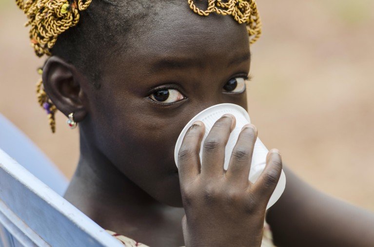 A young African girl looks directly into the lens as she drinks a cup of water