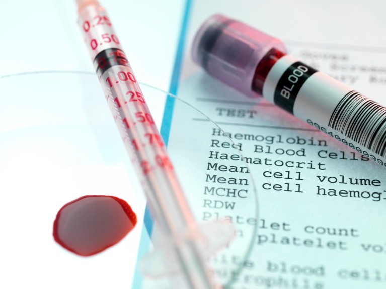 Small pool of blood next to a syringe. Behind is a vial of blood on a printed blood test form.