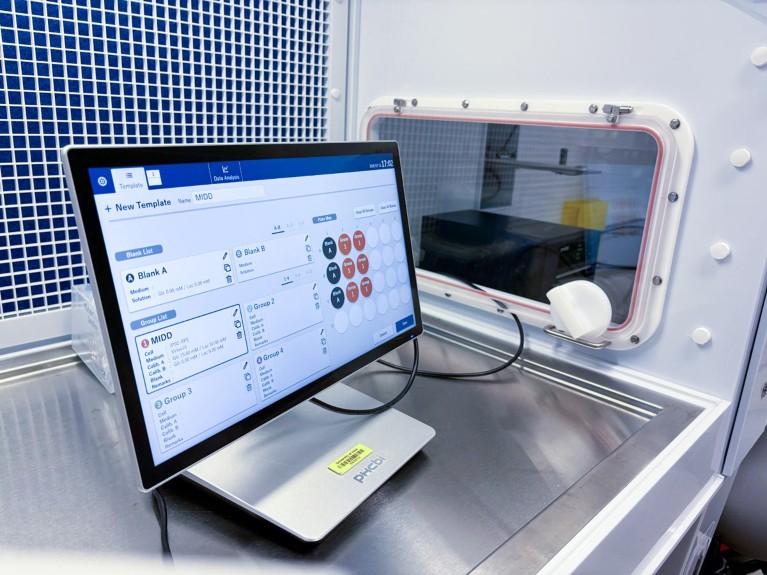 Estación de trabajo informática dentro de una sala limpia de laboratorio o cabina de bioseguridad, equipada con un monitor táctil que ejecuta un software de gestión experimental y conectado por cable a un equipo automatizado protegido por una ventana de visualización sellada. Las superficies de trabajo de acero inoxidable, un panel de ventilación azul con rejilla y un entorno estéril cerrado enfatizan un entorno de laboratorio controlado para cultivos celulares o flujos de trabajo analíticos.