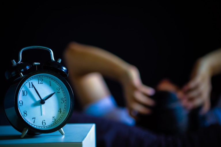 L’image montre une personne qui a du mal à dormir. Une horloge affiche 2h du matin.