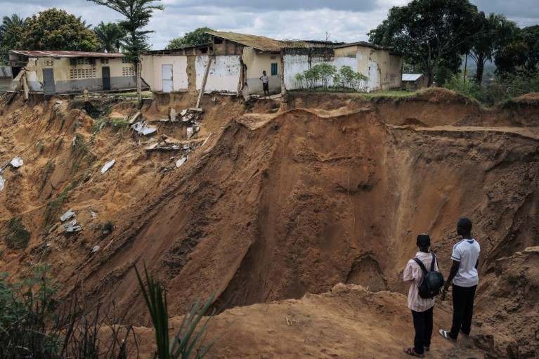 Picture shows two people standing on the edge of a huge sinkhole. On the other side some houses are perched precariously