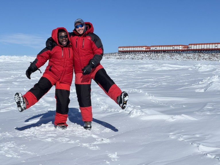 Deux étudiants vêtus de combinaisons polaires rouges prennent une pose enjouée dans un paysage antarctique enneigé. La base de recherche SANAE IV apparaît à l’arrière-plan, sous un ciel bleu dégagé.