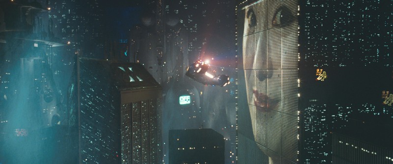 In a futuristic cityscape, a vehicle hovers in front of a screen showing a woman's face.