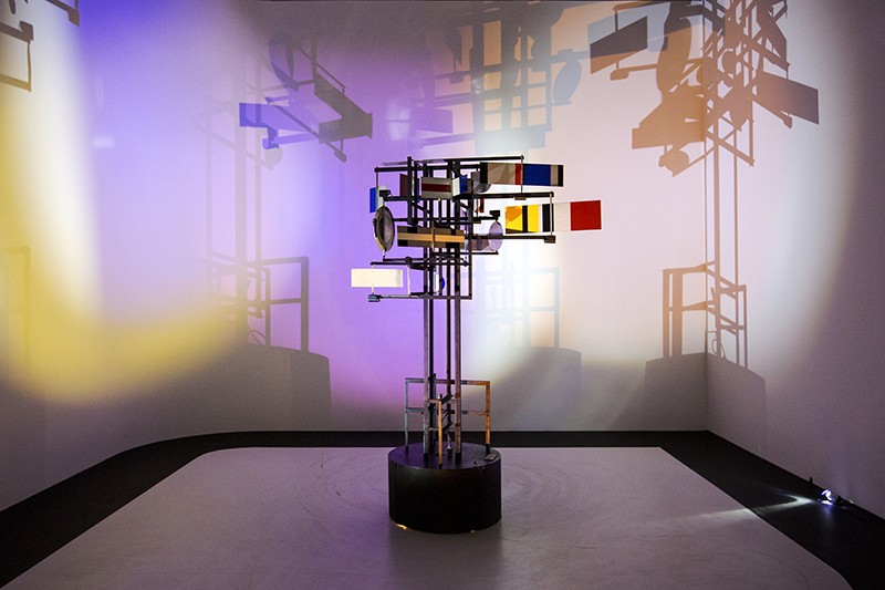 A sculpture made of metal rods and coloured panels stands on a black podium, its shadow cast on the walls in different colours.