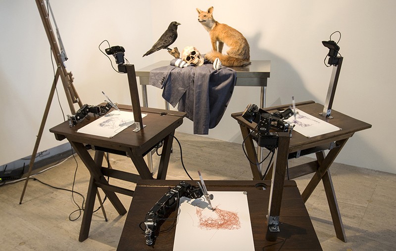 Three sets of robot arms and cameras on tables, drawing a still life of a stuffed fox, a stuffed raven and a human skull.