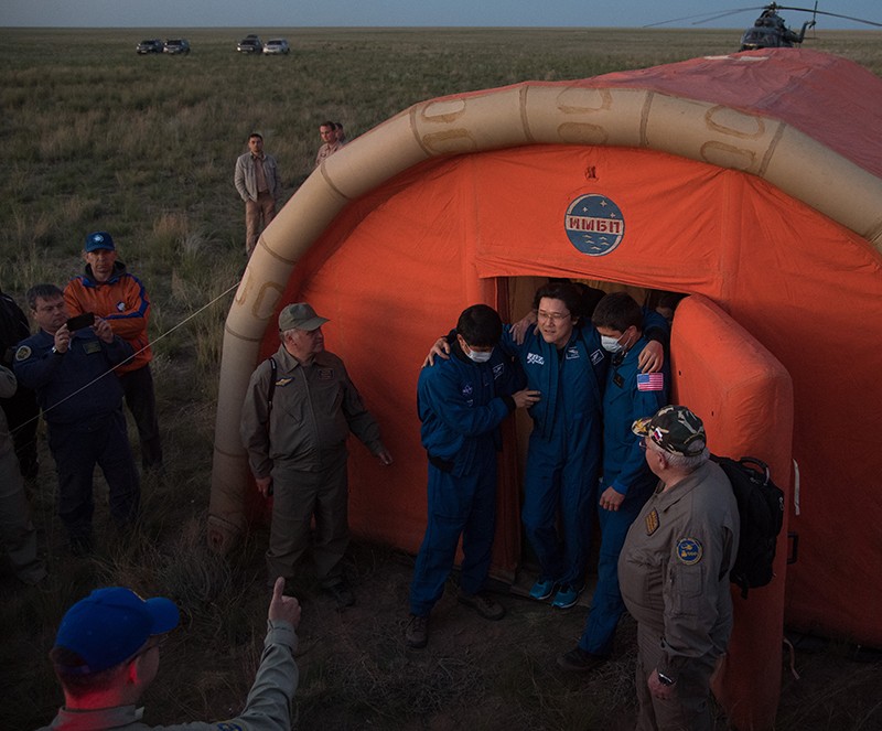 JAXA astronaut Norishige Kanai is helped out of an orange medical tent with grass billowing in the field behind them.