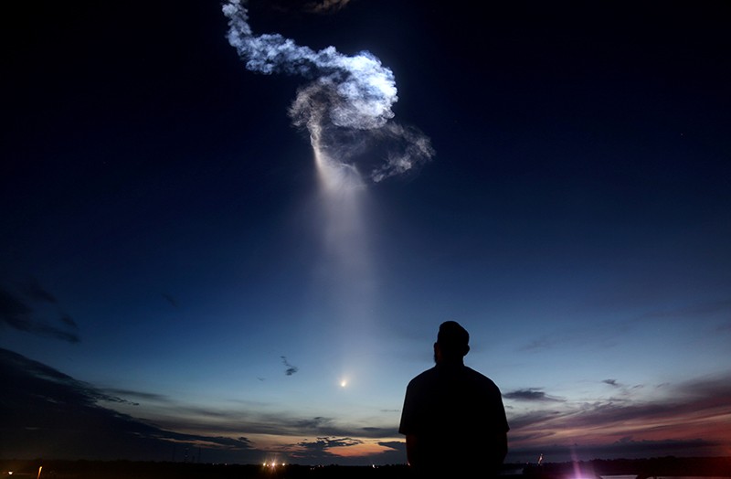 A figure gazes up at the plume of a SpaceX Falcon 9 rocket exiting the atmosphere during a sunset.