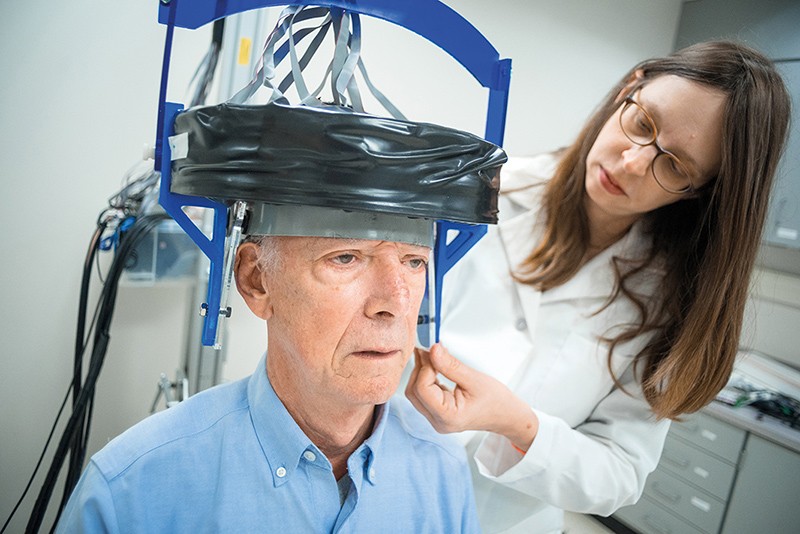 A researcher adjusts a prototype head-mounted positron emission tomography scanner worn by a male volunteer.