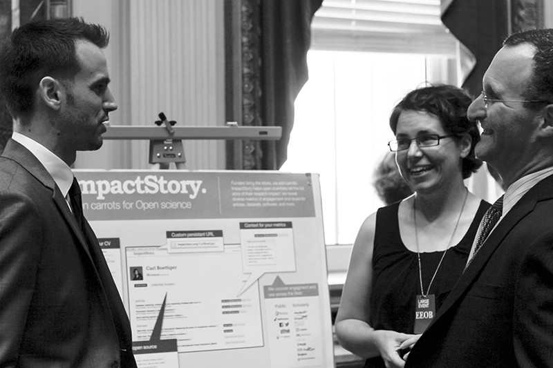 B+W photo of Priem (L) and Piwowar (c) talking to Rossner (r) in front of an ImpactStory presentation board at the White House