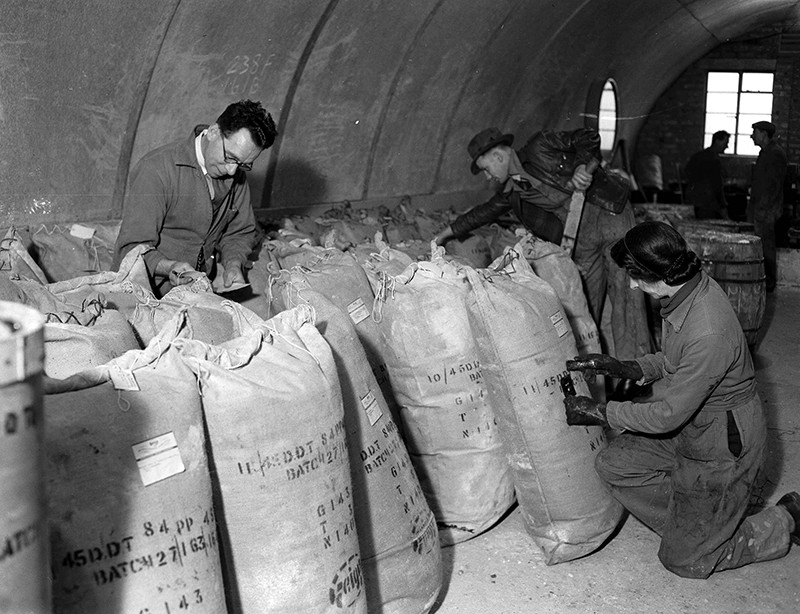Trei persoane împachetează DDT brut în saci într-un hangar/bunker.