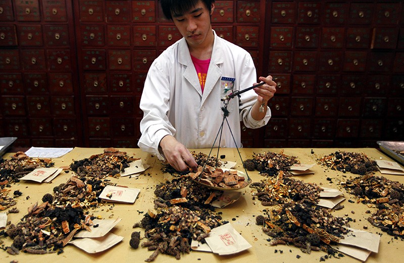 A traditional-medicine pharmacist stands behind a desk covered in dried product