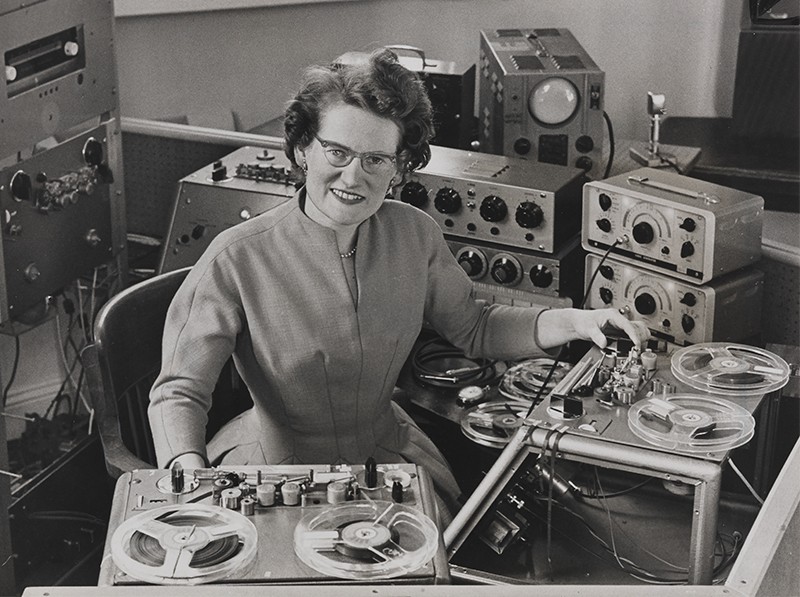 Daphne Oram smiles in her Kent studio surrounded by tape machines, mixers and electronic equipment.