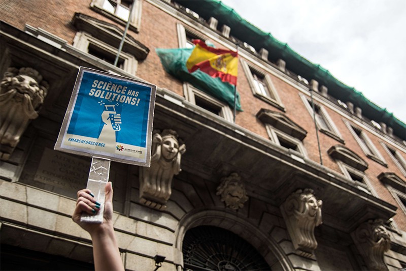A woman holds up a ‘Science has Solutions’ sign during the March for Science, April 2017, with buildings and flags in background